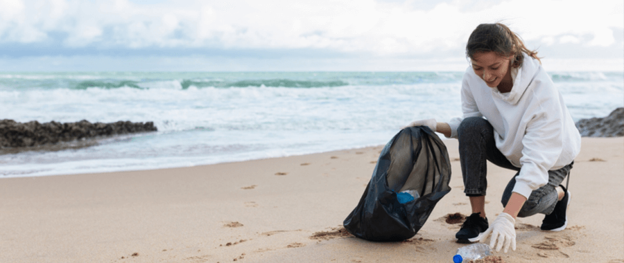 A woman on the beach picking up trash and placing it in a bag.